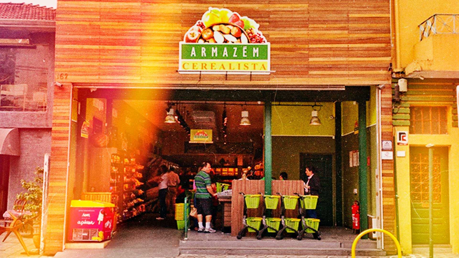 Fachada frontal da loja Armazém Cerealista, com uma placa acima da entrada com frutas na parte superior e as escritas Armazém em um retângulo verde a baixo das frutas e Cerealista em outro retângulo na cor amarela a baixo.
